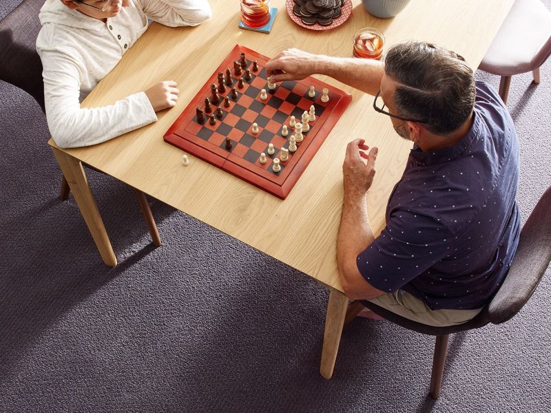 two men playing chess at a wooden table in a room with purple carpet from Northcraft Flooring & Design in Lee's Summit, MO